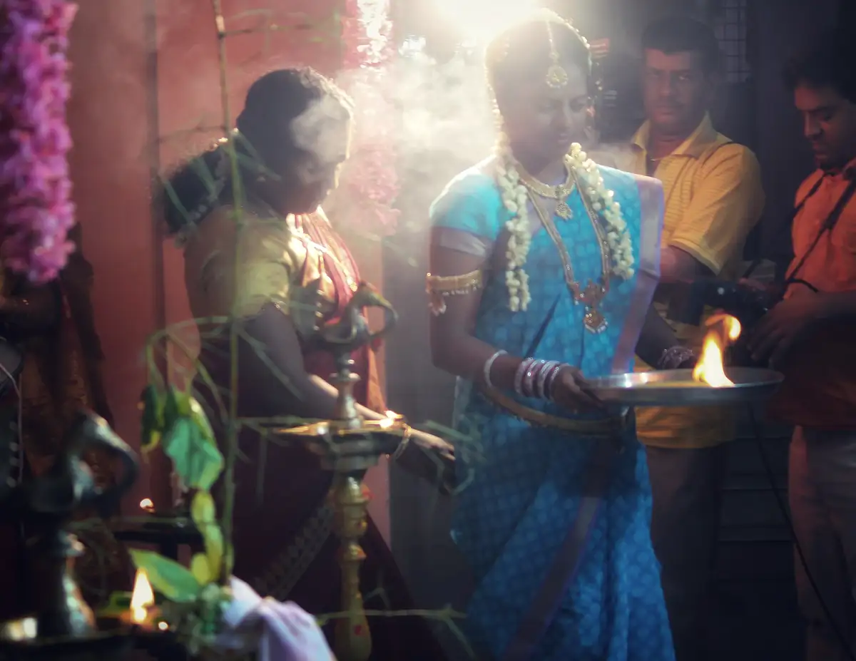 A bride carries an aarti plate through camphor smoke and a shaft of light beside a brass lamp